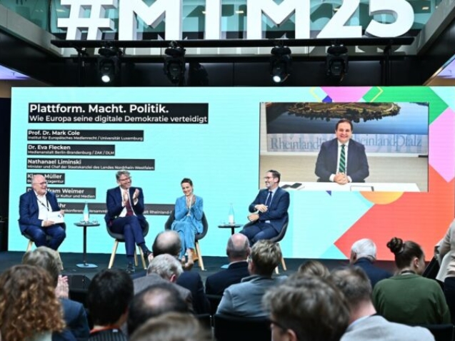 Podiumsdiskussion mit Experten und Politikern zum Thema 'Plattform. Macht. Politik. Wie Europa seine digitale Demokratie verteidigt'. Im Hintergrund eine große Leinwand mit Livestream, im Vordergrund ein interessiertes Publikum.